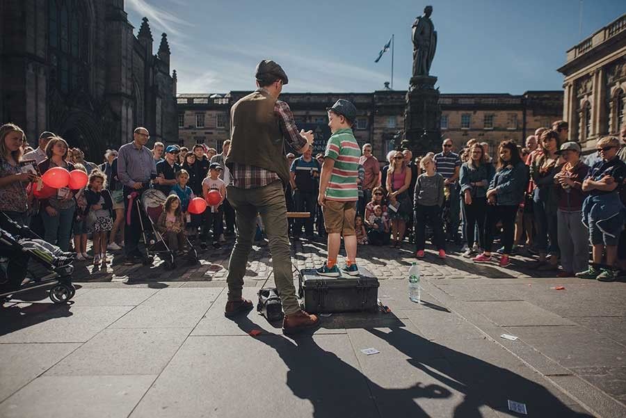 A street performer and a young boy standing on a brown suitcase on the Royal Mile, at the Edinburgh Festival.