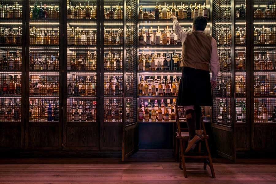 A man in a kilt gets a whisky bottle from a large whisky shelf.