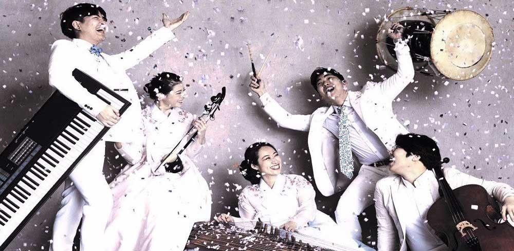 A group of musicians dressed in white, standing in various poses with white confetti falling around them.